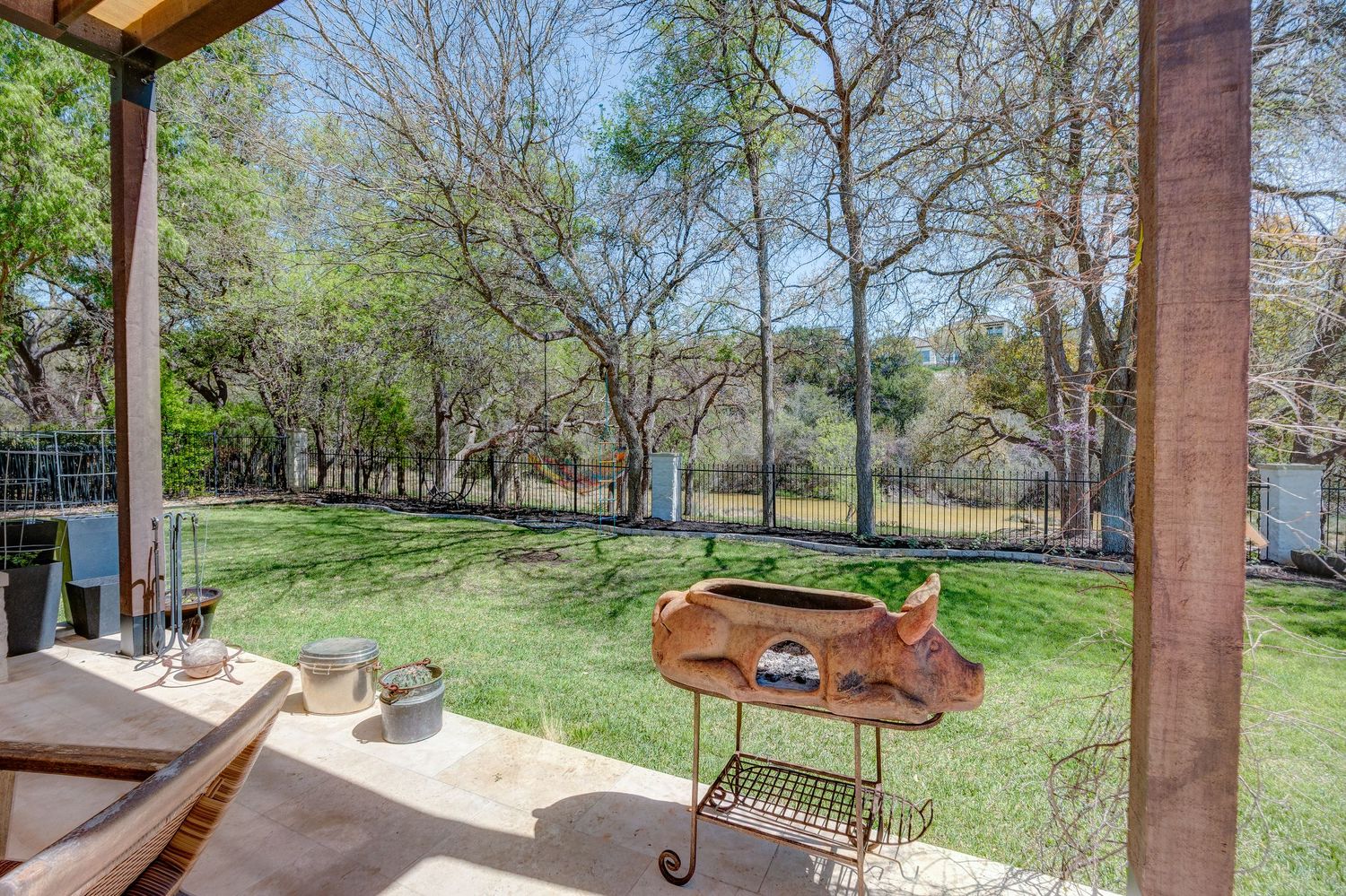 Covered patio with decorative metal pig sculpture overlooking a lush green backyard with mature trees and fenced yard.