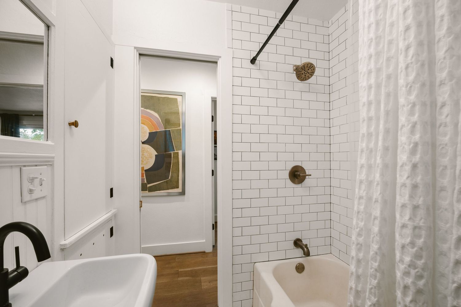 White subway tile bathroom with clawfoot tub, brass fixtures, white curtain, and stained glass window visible through doorway.