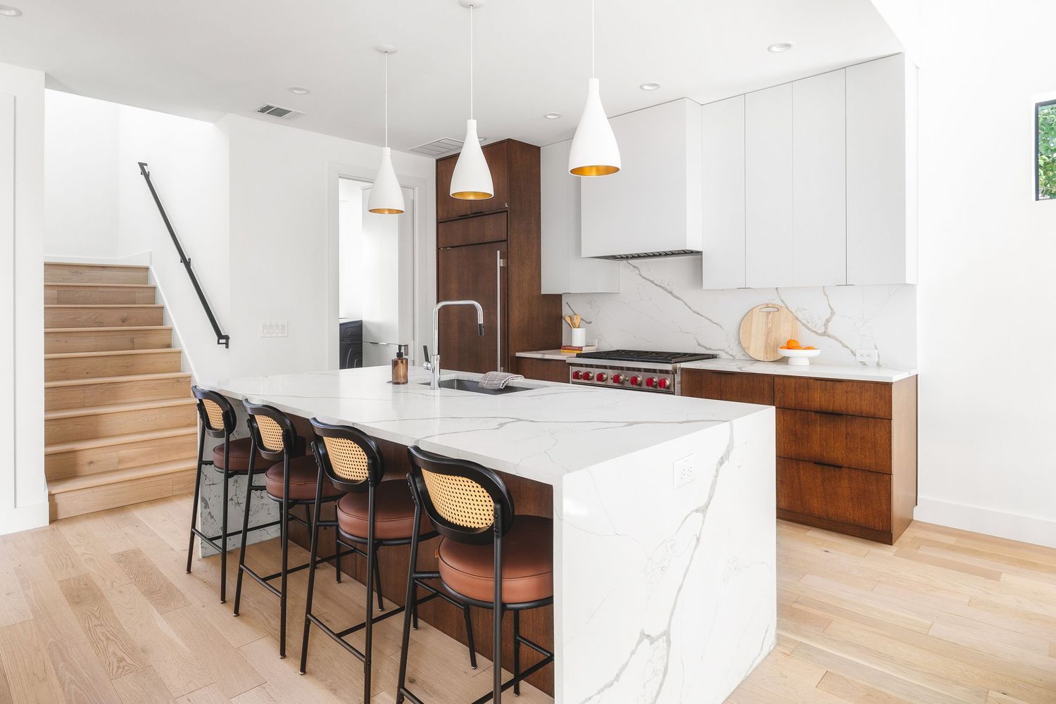 Modern white kitchen with marble island, wooden cabinets, pendant lights and bar seating overlooking staircase.