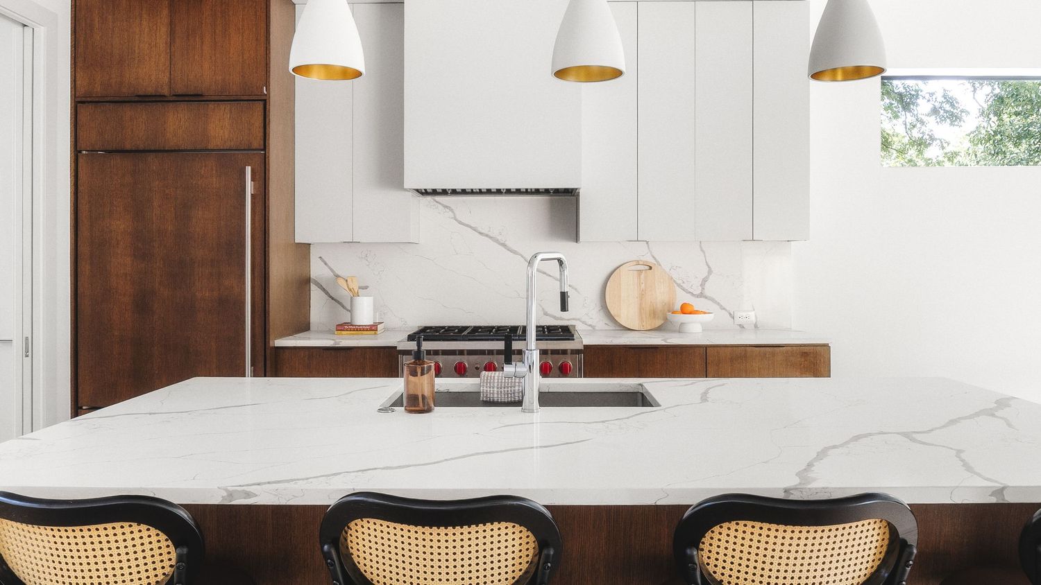 Modern white kitchen with wood accents features pendant lights and rattan counter stools at a large marble island.