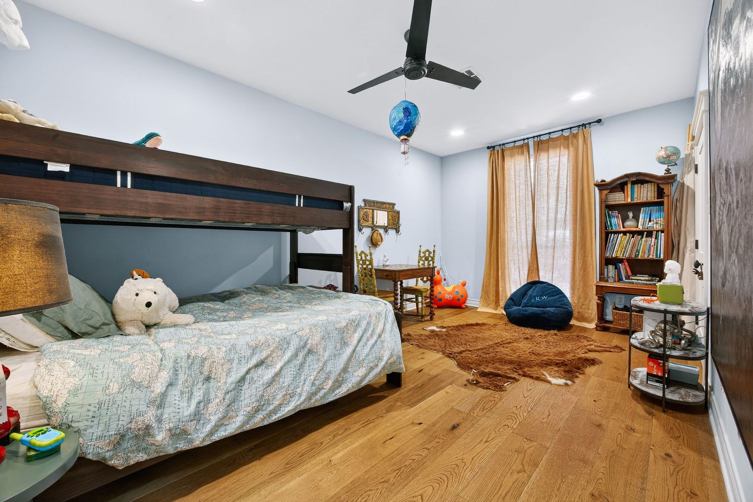 Children's bedroom with bunk beds, hardwood floors, bookshelf, bean bag chair, and warm curtains lit by recessed lighting.