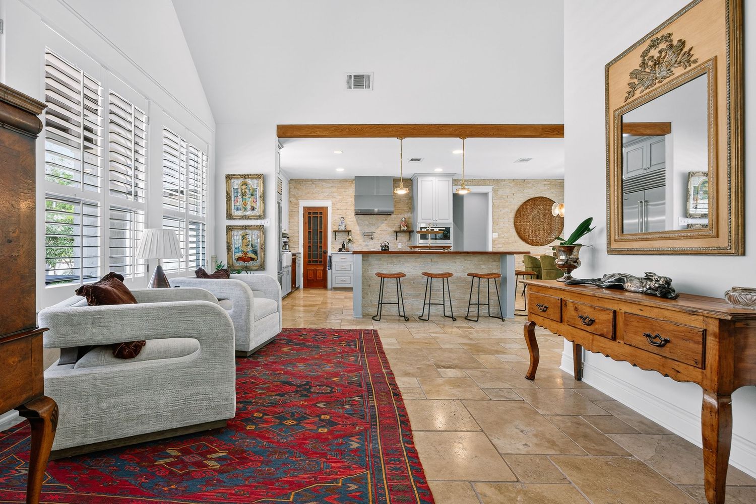 Bright open living room with red Persian rug, tile floors, plantation shutters, and view of modern kitchen with bar seating.