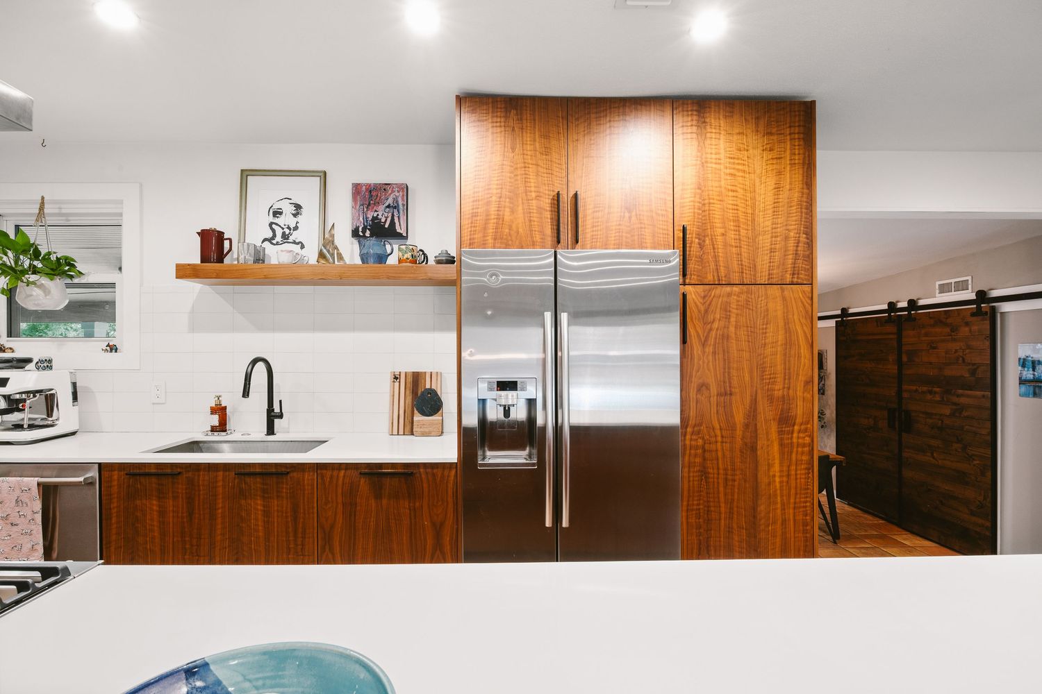 Modern kitchen with walnut cabinets, stainless steel fridge, floating shelf, and black sink in a bright white space.