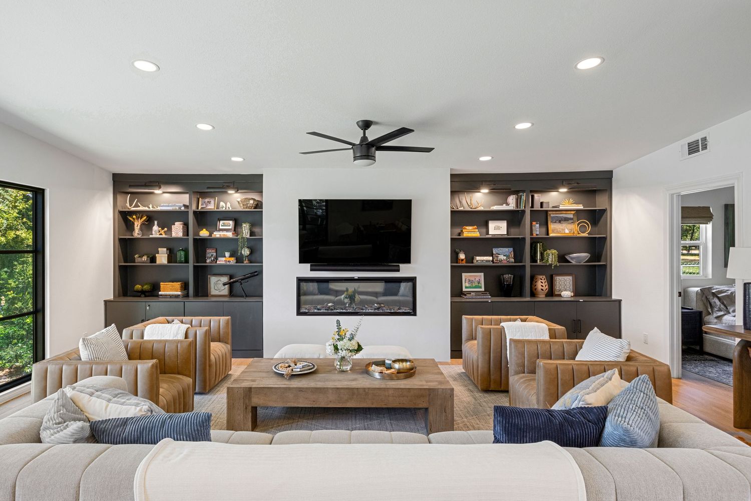 Elegant living room with custom built-in shelving, wall-mounted TV, fireplace, ceiling fan, and plush neutral sectional sofa.