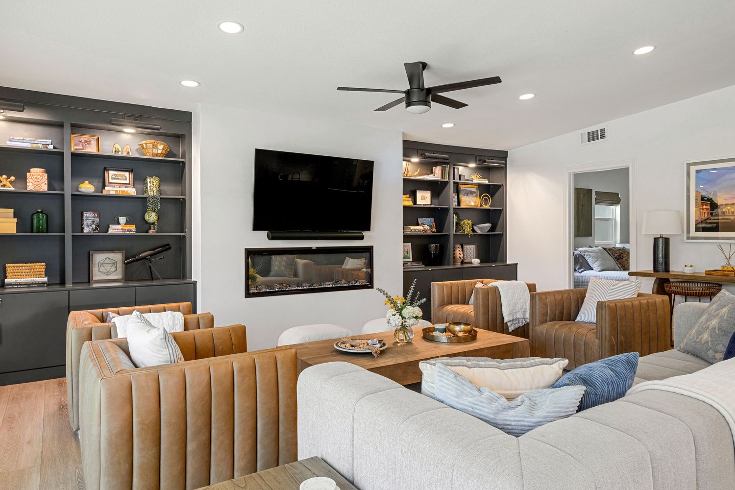 Modern living room with built-in shelves, wall-mounted TV, linear fireplace, ceiling fan, and tan leather chairs.