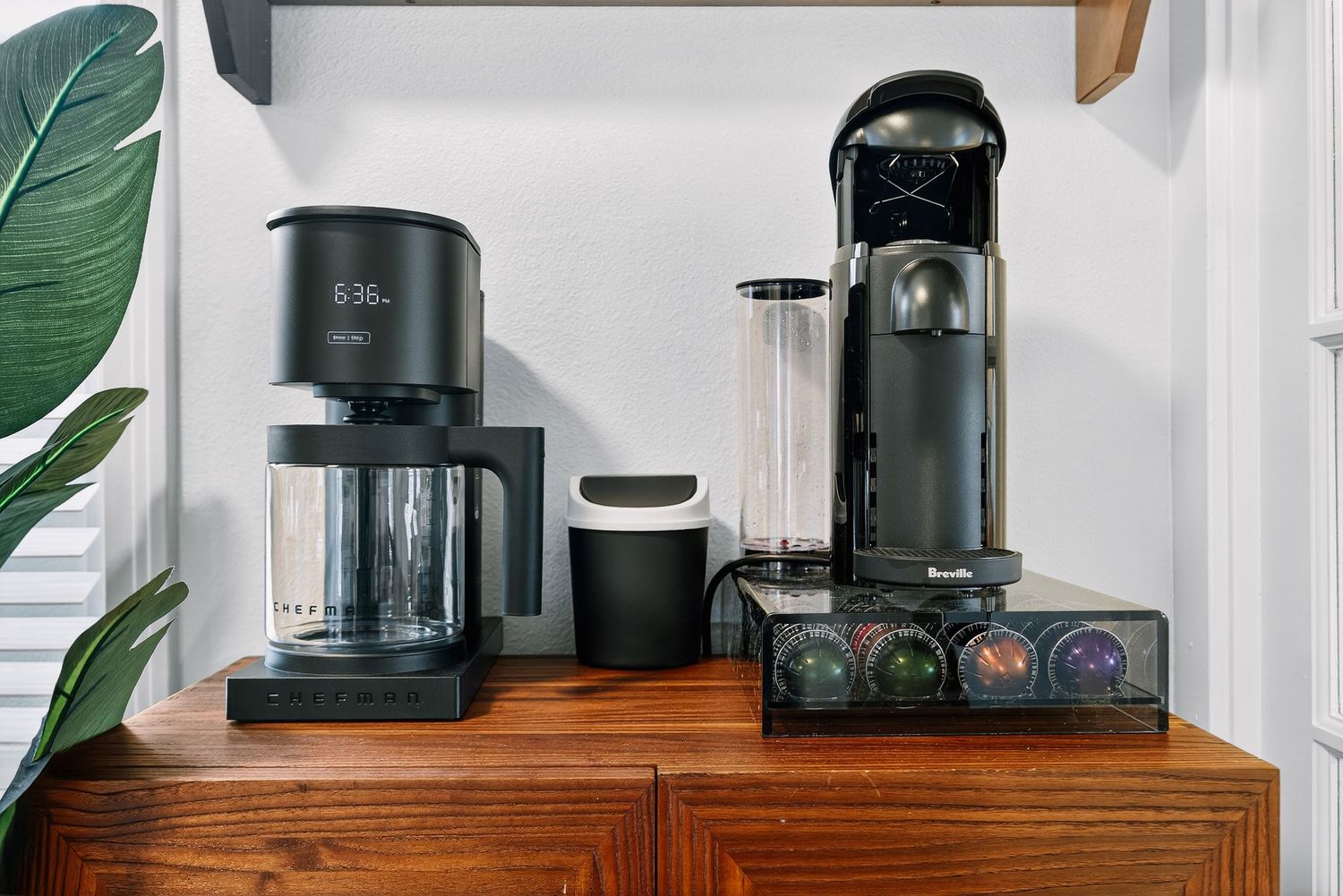 Modern coffee station setup on wooden cabinet with black coffee makers and green plant decor against white wall.