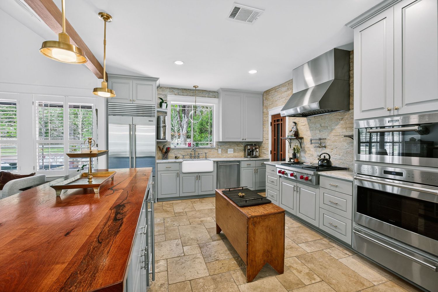 Modern kitchen with gray cabinets, wood island, stainless appliances, stone backsplash, and pendant lights over butcher block counter.