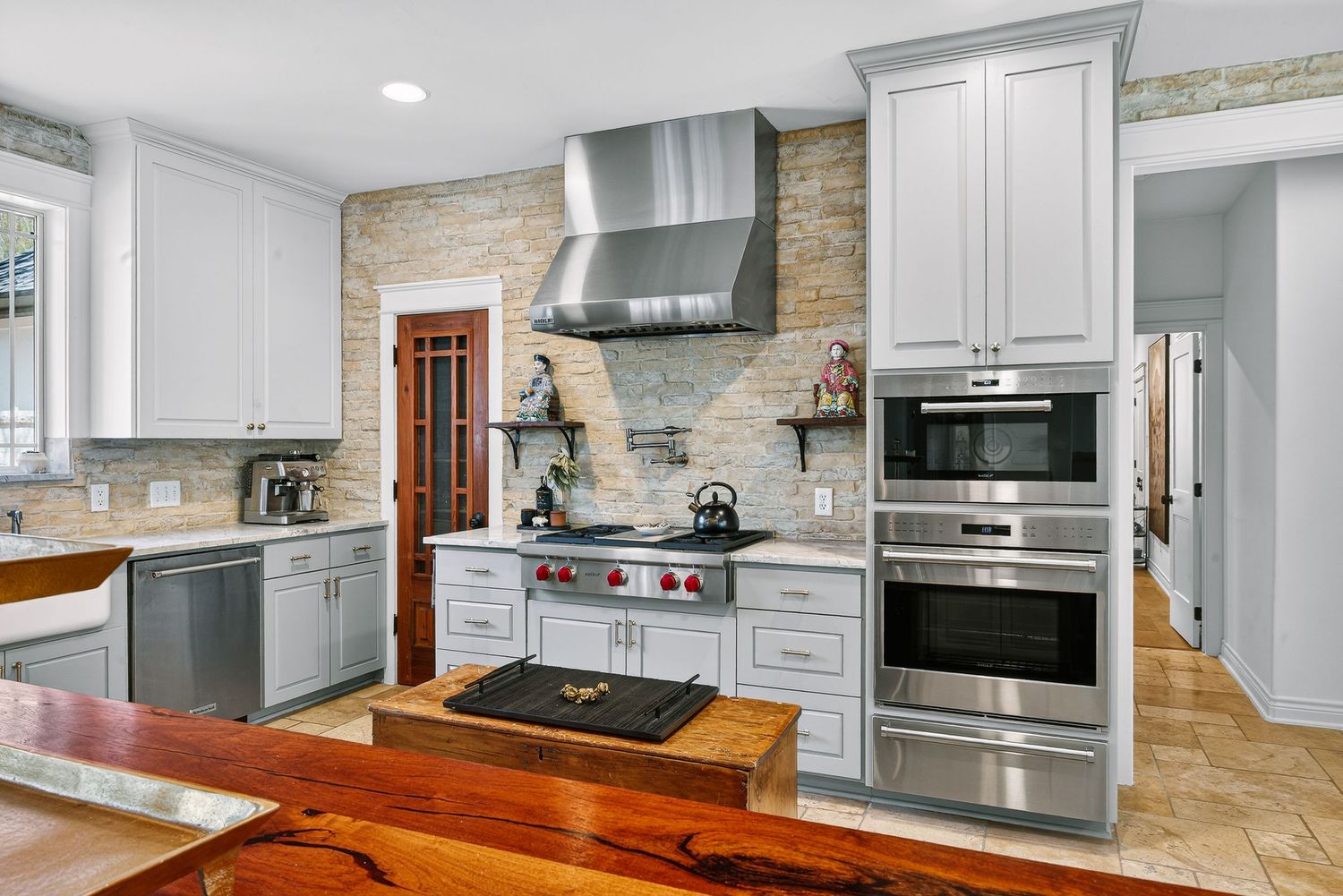 Modern kitchen with white cabinets, stainless steel appliances, stone backsplash, range hood, and rustic wood countertop accents.