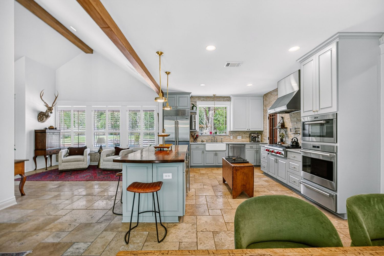 Open-concept kitchen with gray cabinets, island seating, stainless appliances, exposed wood beams, and tile flooring in a bright home.