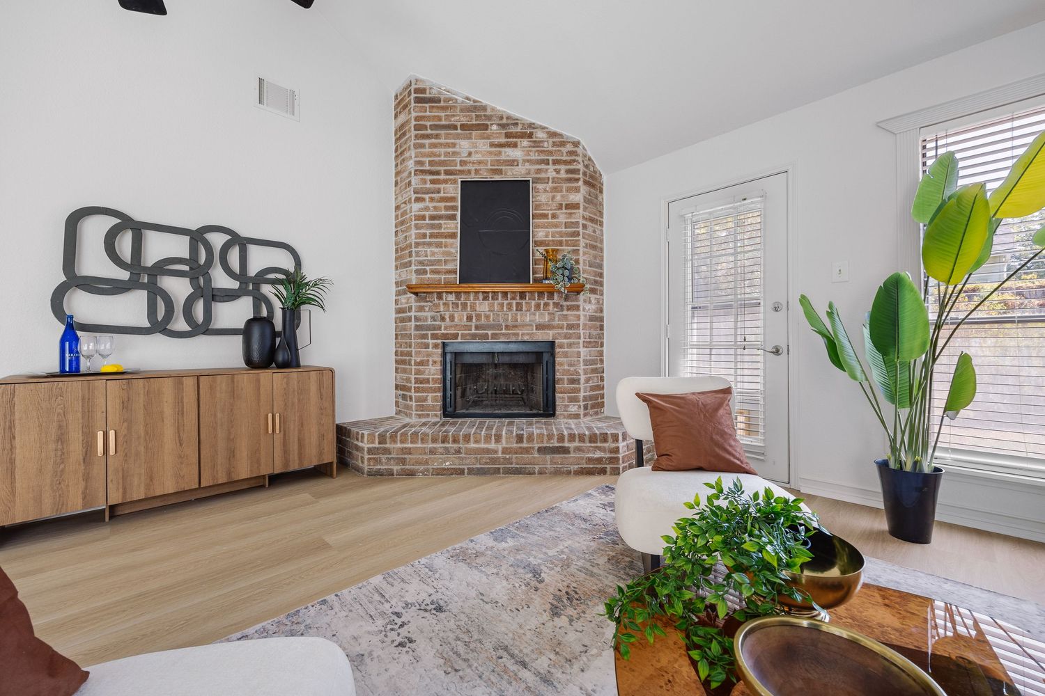 Bright living room with a vaulted ceiling, brick fireplace, light hardwood floors, wood sideboard, accent chair, and leafy green plants.