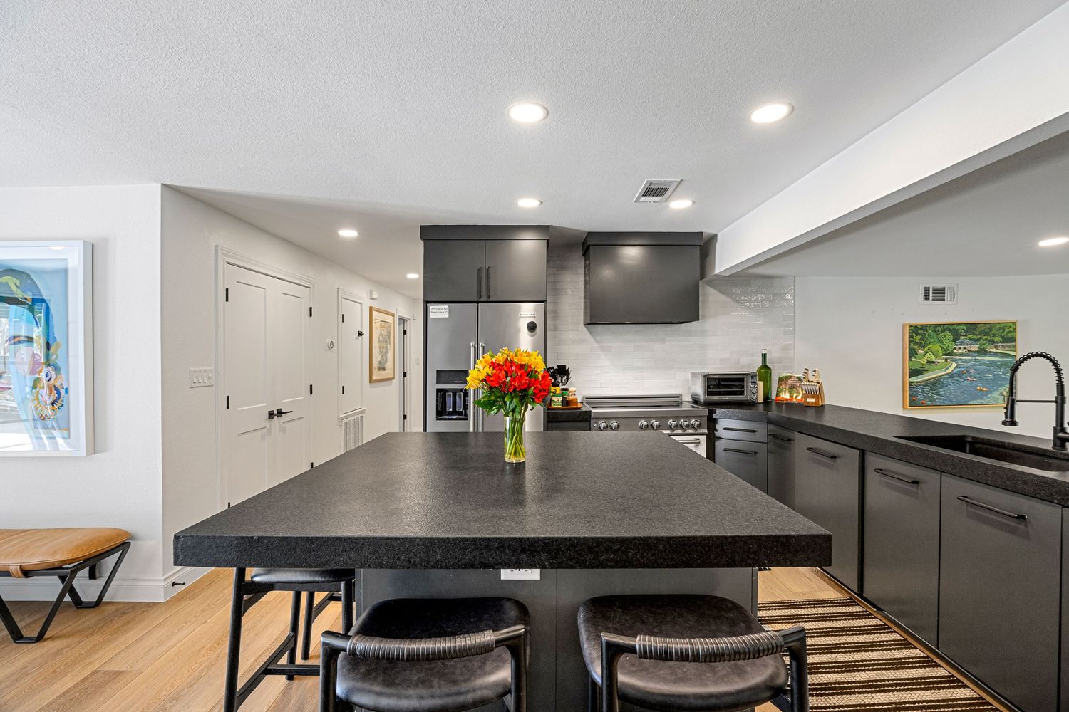 Contemporary kitchen featuring black countertop island with bar seating, dark cabinetry, stainless steel appliances, and hardwood floors.