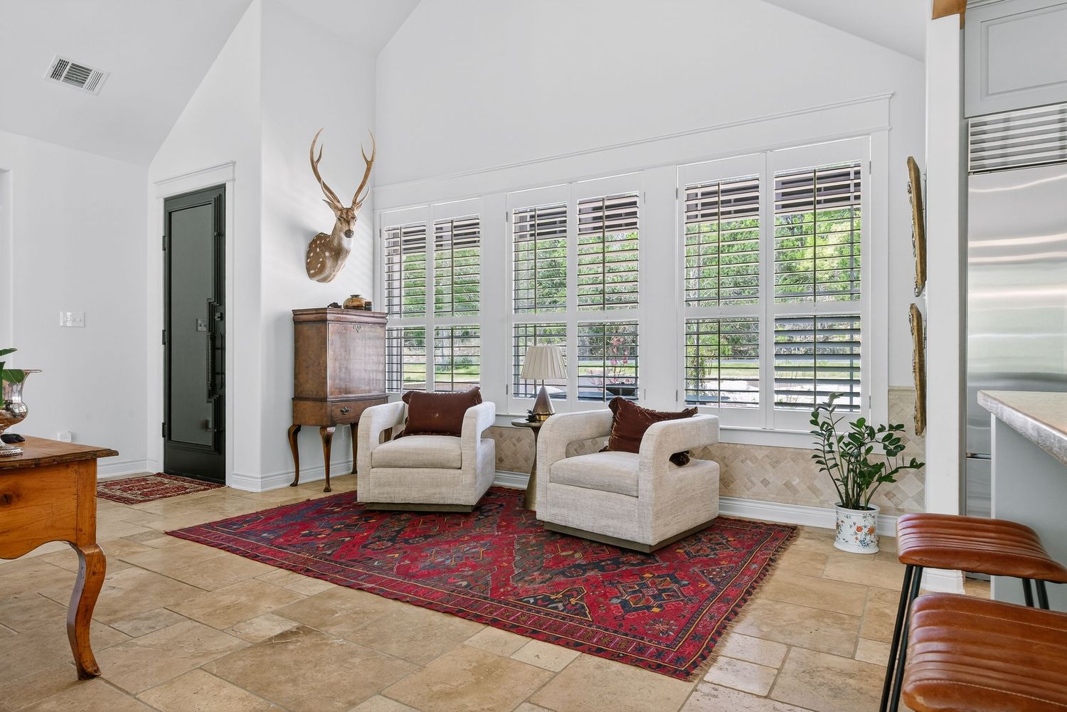 Bright living room with vaulted ceilings, white plantation shutters, two cream armchairs, a red Persian rug, and mounted antler decor.