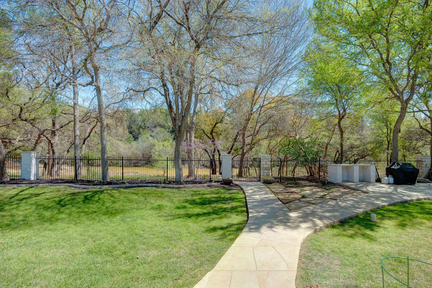 A winding stone pathway leads through a lush green lawn surrounded by tall bare trees in a serene backyard garden.