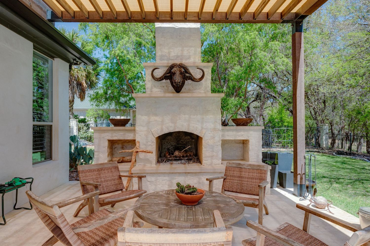 Outdoor patio with stone fireplace, teak chairs, round table, and buffalo mount under a wooden pergola surrounded by trees.