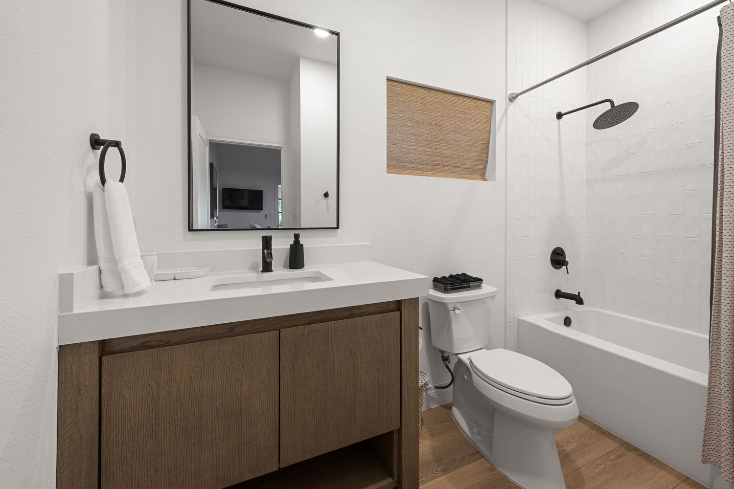 Clean bathroom with wood vanity, black framed mirror, white countertop, toilet, and tub-shower combo with rain head.