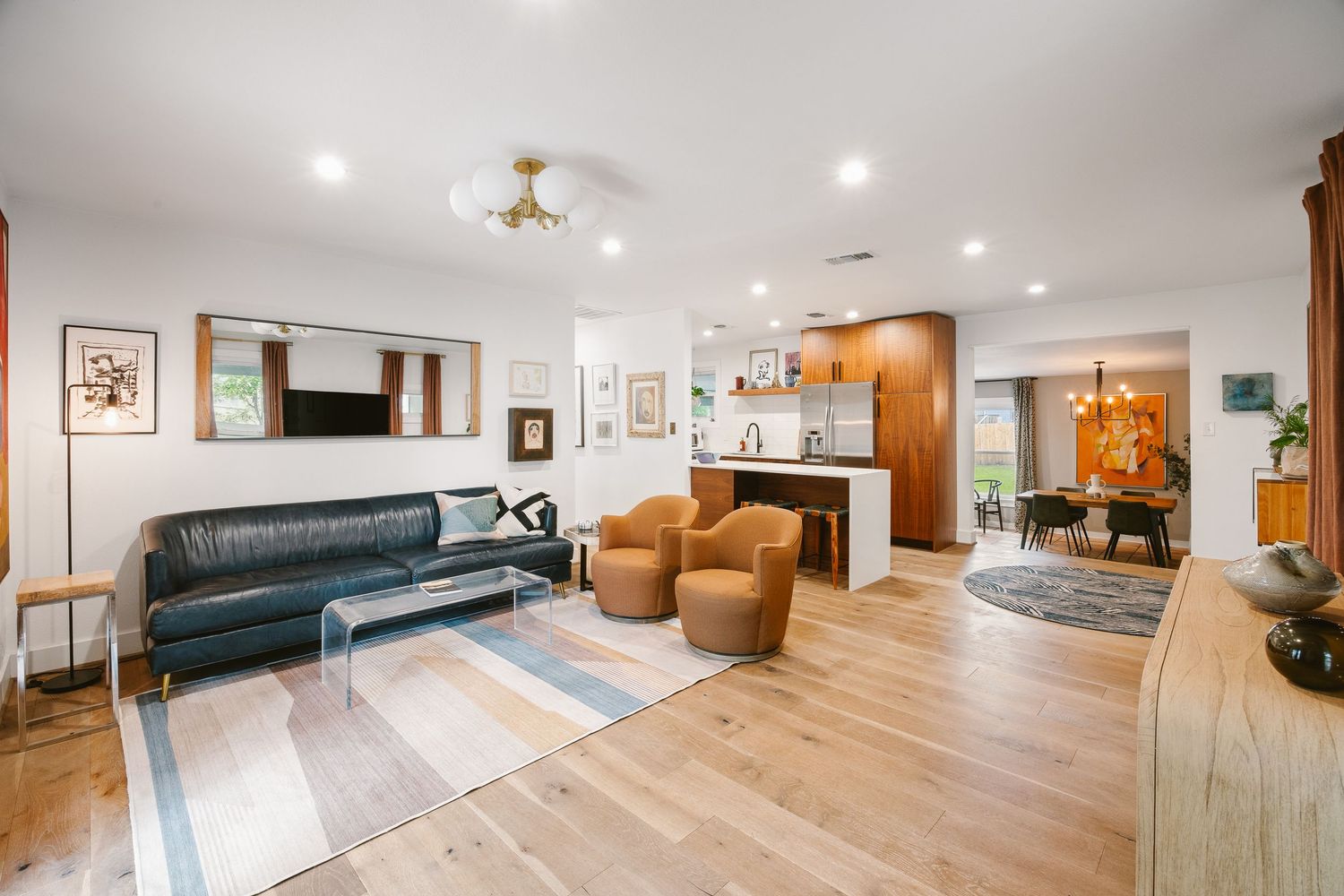 Modern open-plan living room with leather sofa, hardwood floors, striped rug, and open kitchen in background.