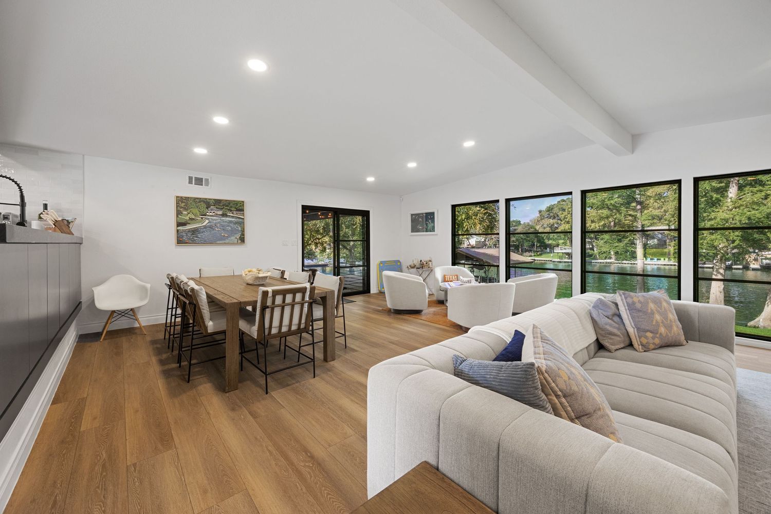 Bright open-plan living and dining area with beamed ceiling, hardwood floors, large windows, and modern neutral furnishings.