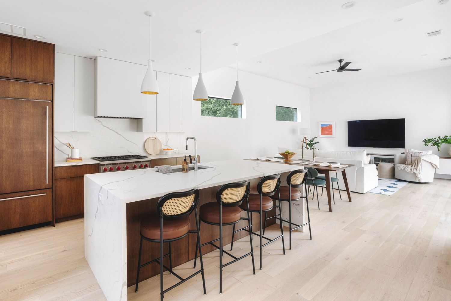 Modern white kitchen with waterfall island, black bar stools, and warm wood cabinets in an open concept space.
