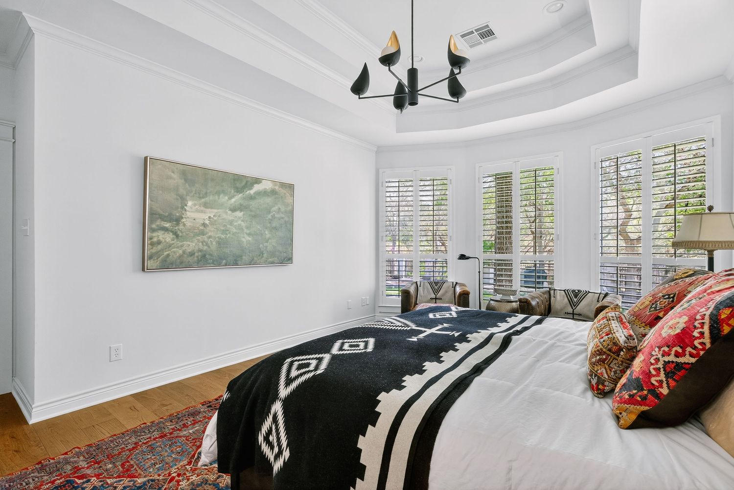 Elegant bedroom with tray ceiling, modern chandelier, plantation shutters, hardwood floors, and bold black-and-white bedding on a king bed.