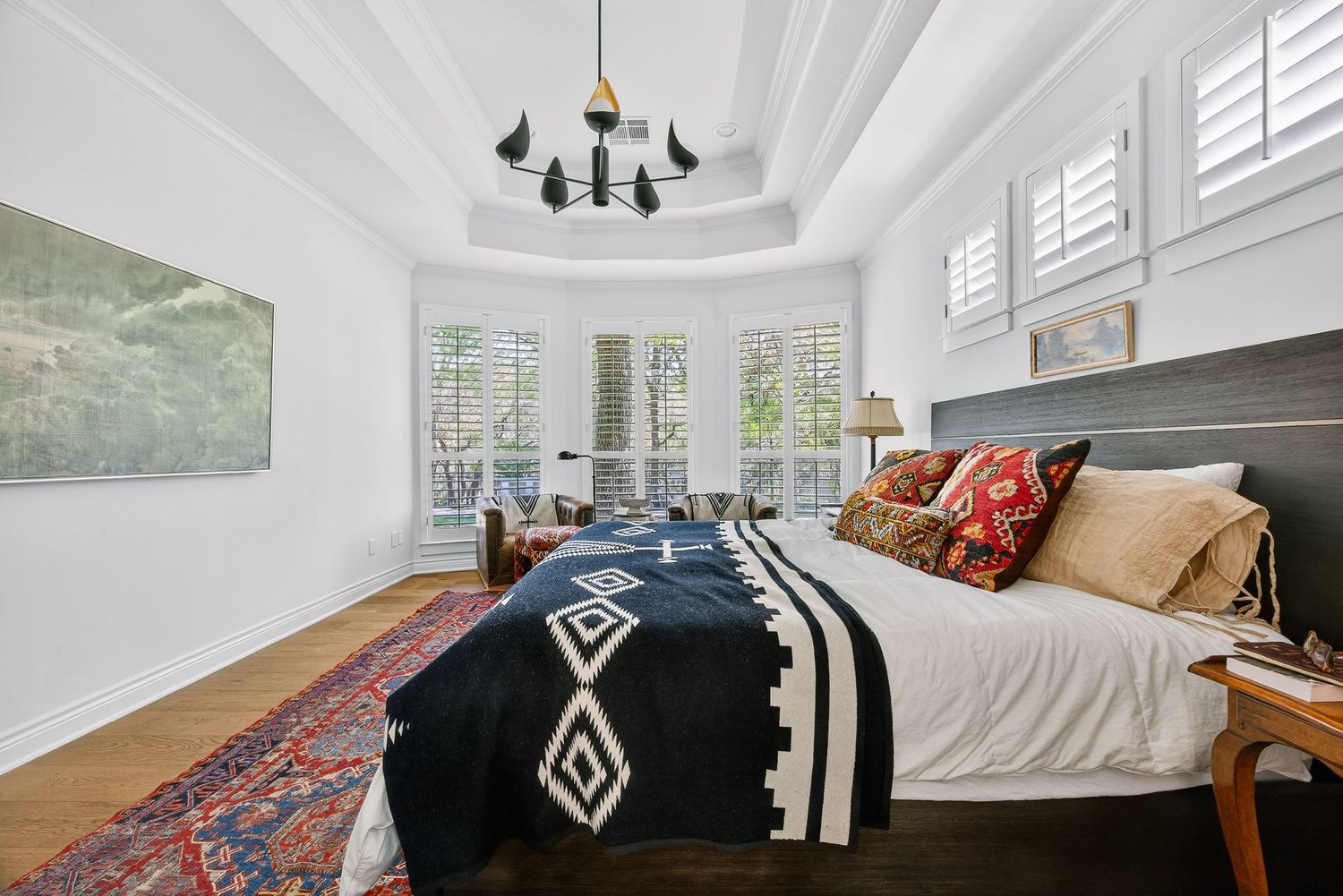 Elegant bedroom with tray ceiling, black chandelier, navy patterned bedding, hardwood floors, and shuttered windows.
