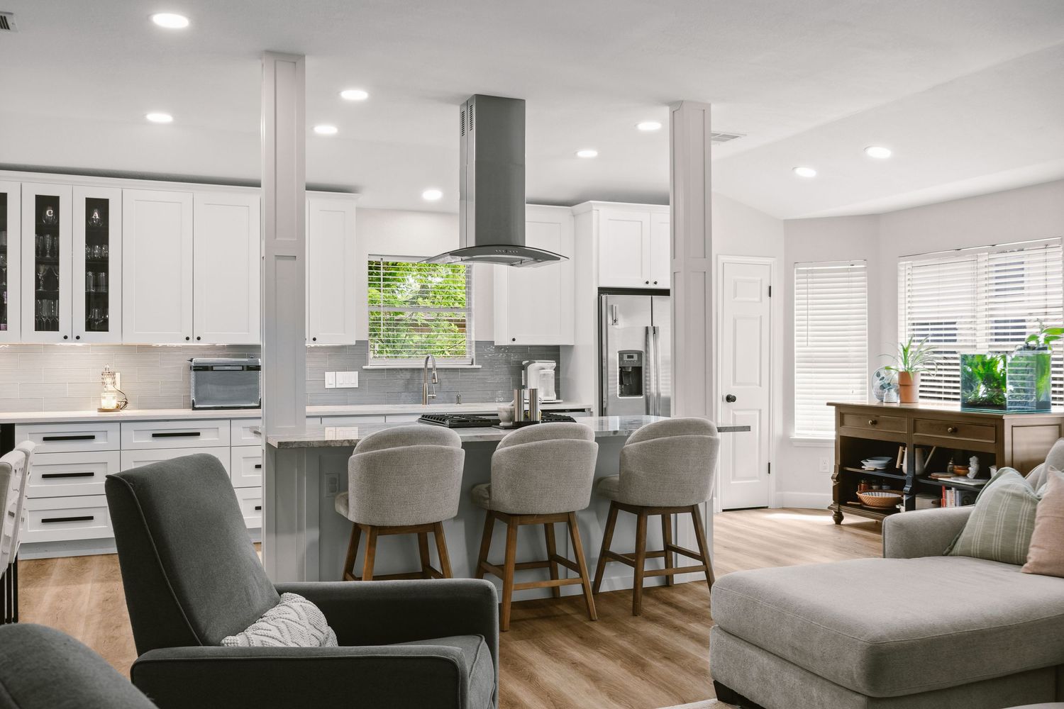 Modern open-concept kitchen with white cabinets, gray island, bar stools, stainless steel appliances, and hardwood floors.