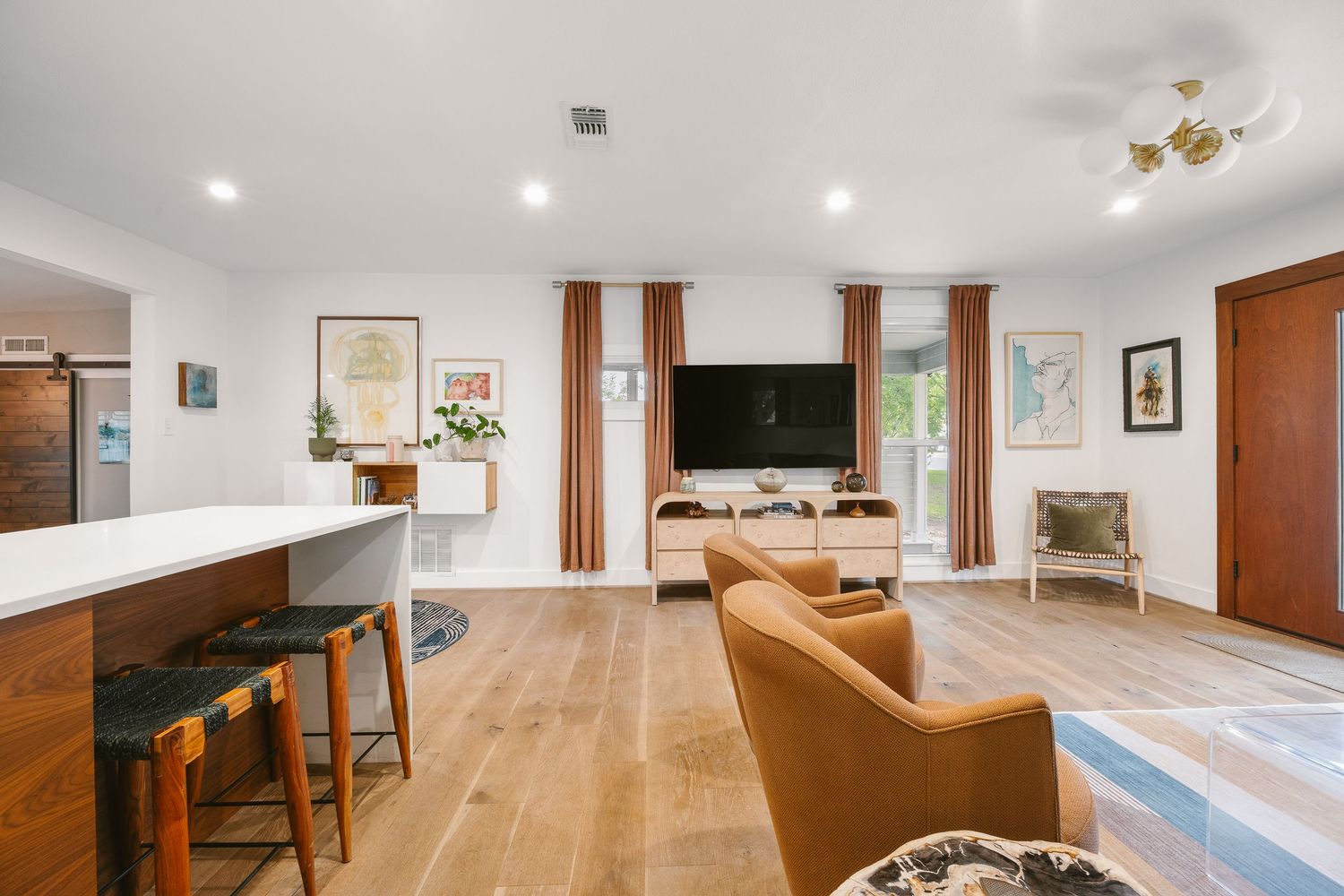 Bright open-concept living room with hardwood floors, leather chairs, wall-mounted TV, and warm natural light through curtained windows.