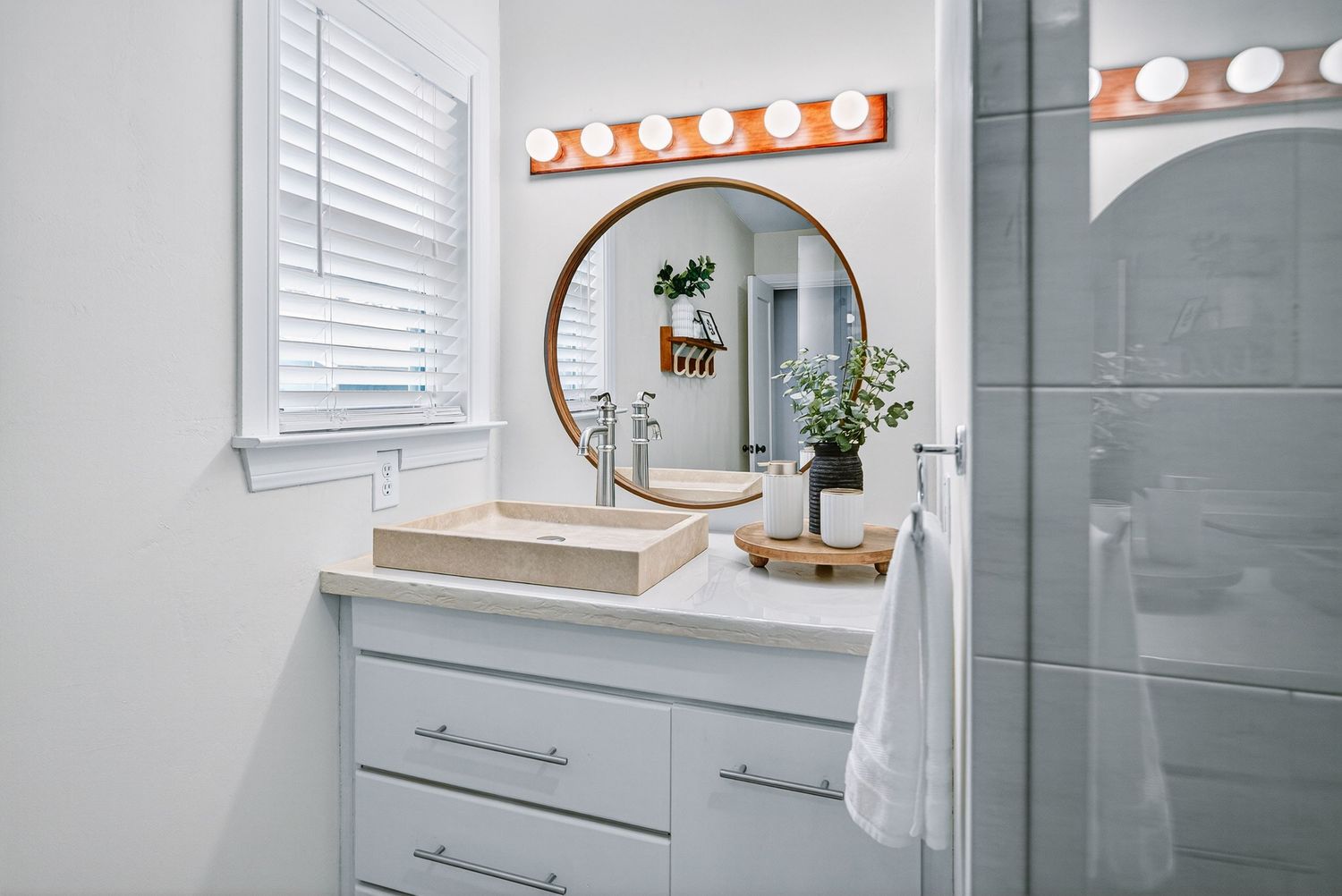 Modern bathroom vanity with round gold mirror, white cabinets, and vintage-style light fixture above the mirror.