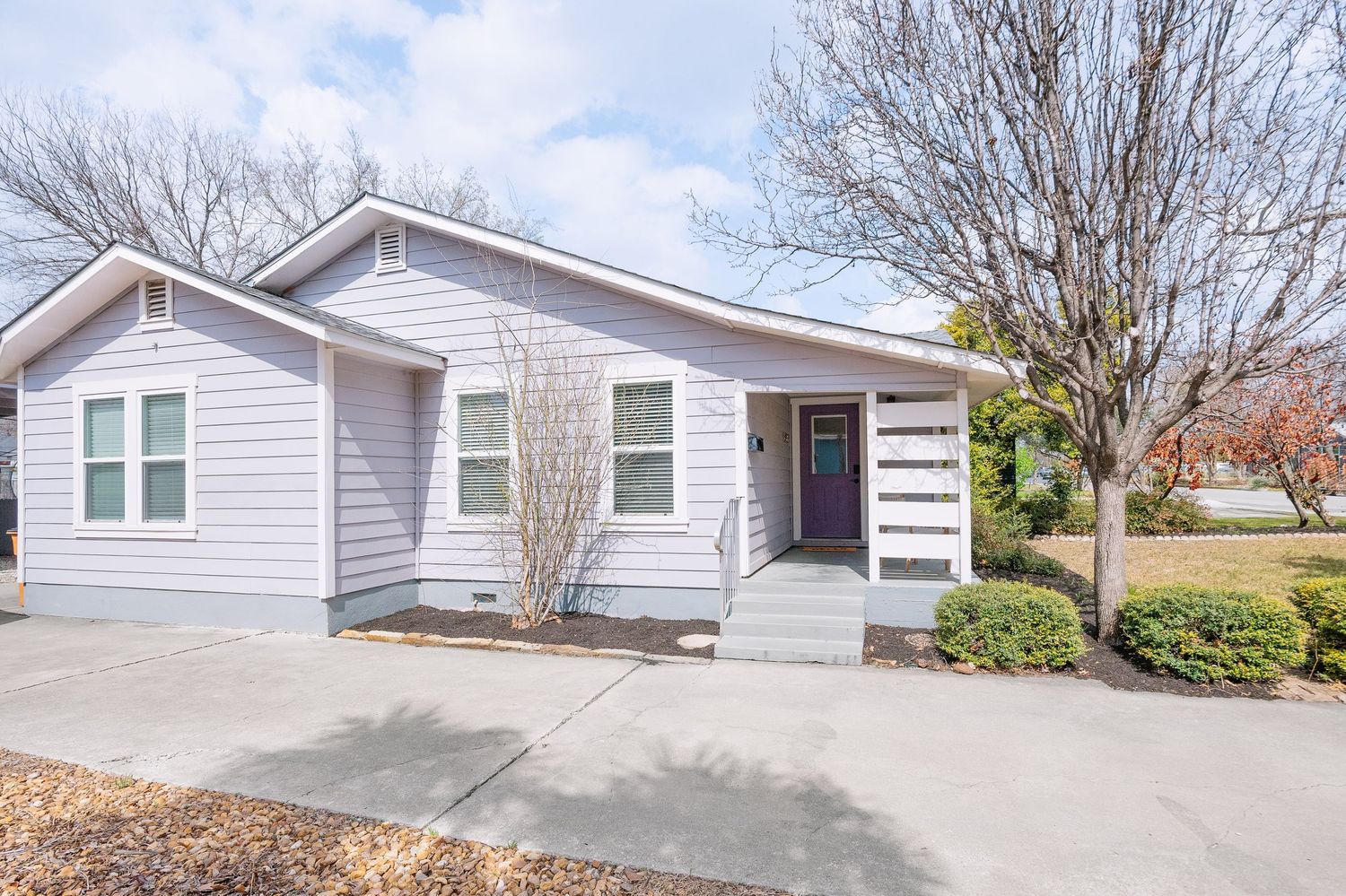 Charming light gray ranch-style home with covered front porch, concrete driveway and landscaped yard on a corner lot.