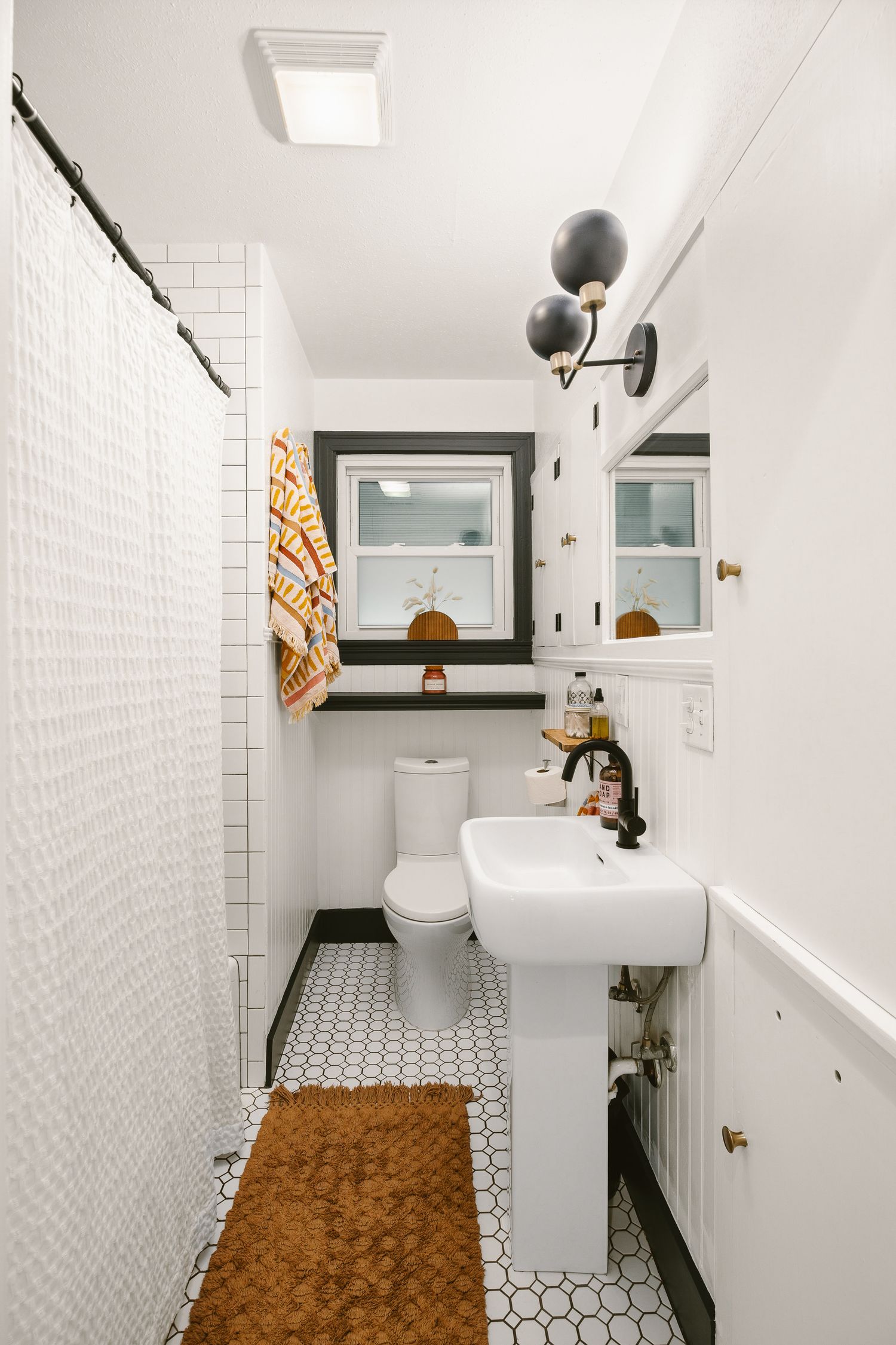 White bathroom with penny tile floor, pedestal sink, black sconces, subway tile shower, and brown bath mat.