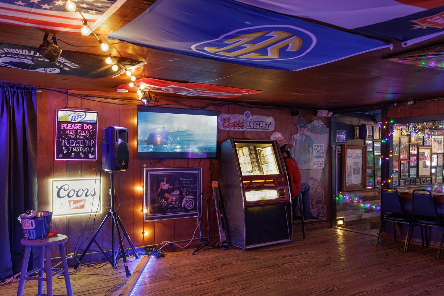 Cozy bar interior with jukebox, Coors neon sign, flat-screen TV, string lights, and hardwood floors.
