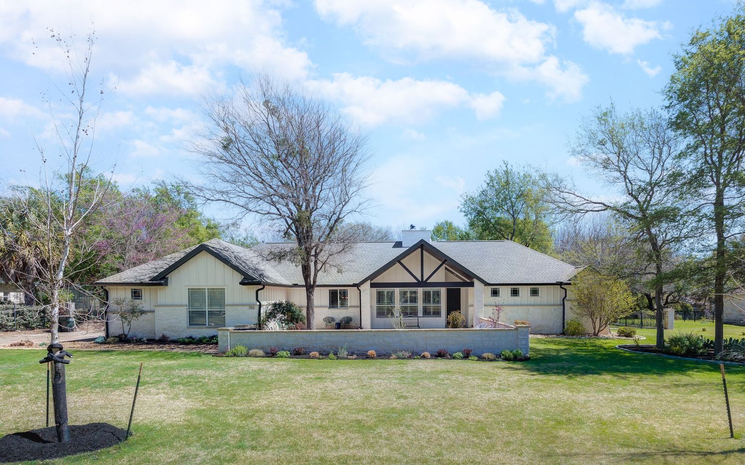 Single-story ranch-style home with white exterior, stone accents, and lush green lawn surrounded by mature trees.