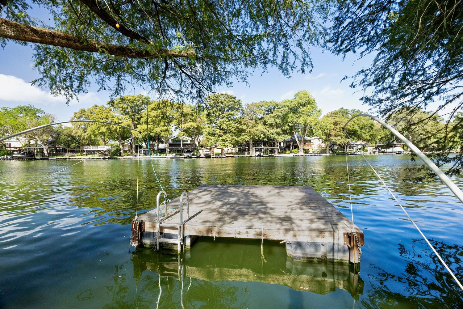 Floating dock on a calm lake surrounded by lush green trees and waterfront homes on a sunny day.