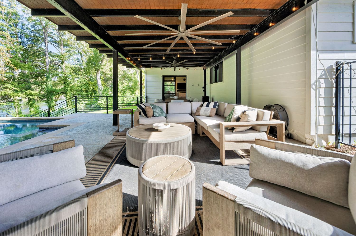 Elegant covered patio with neutral outdoor furniture, string lights, and exposed wood beam ceiling near a pool.