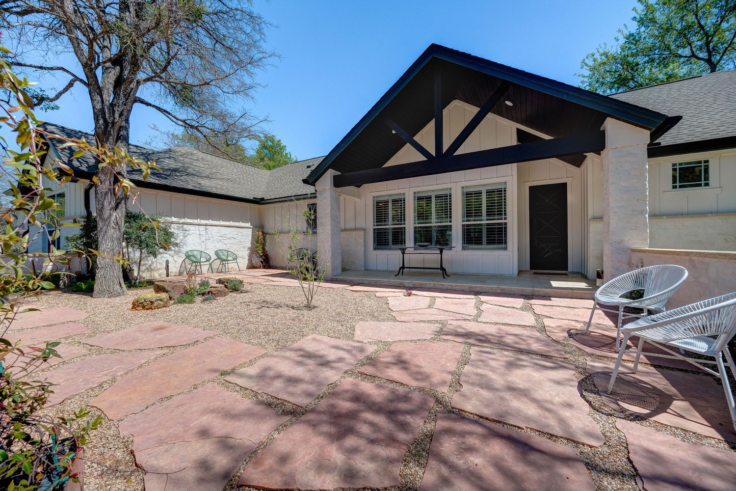 Charming ranch-style home featuring a spacious brick patio, white chairs, and a covered entryway surrounded by mature trees.