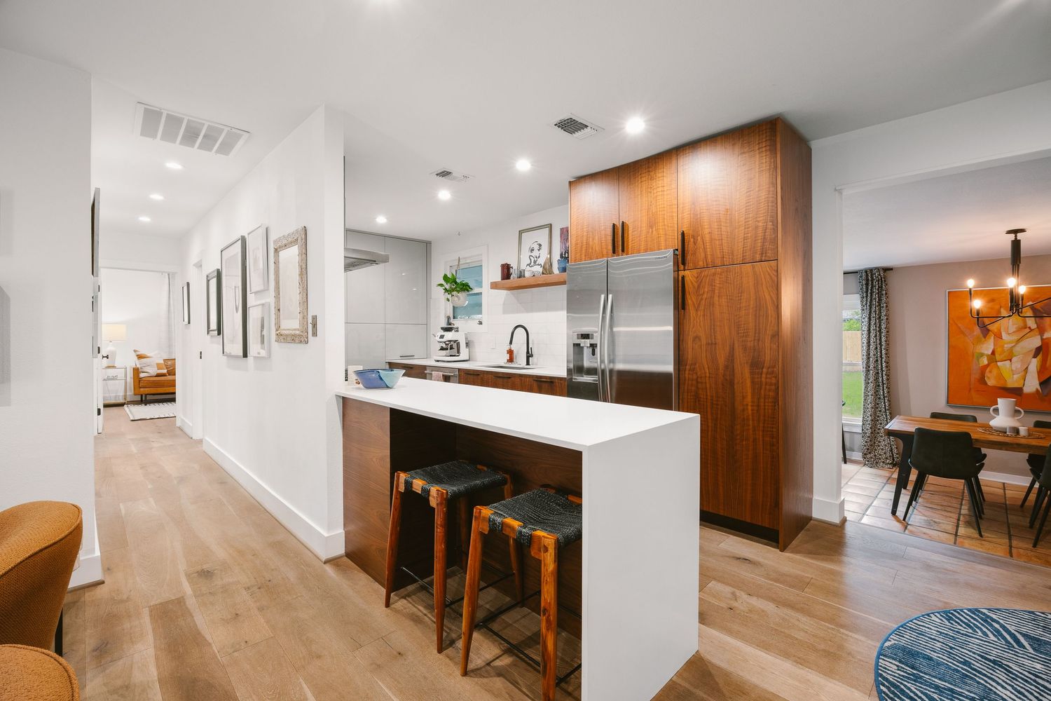 Modern kitchen with white peninsula, bar stools, walnut cabinets, stainless steel fridge, and light hardwood floors.