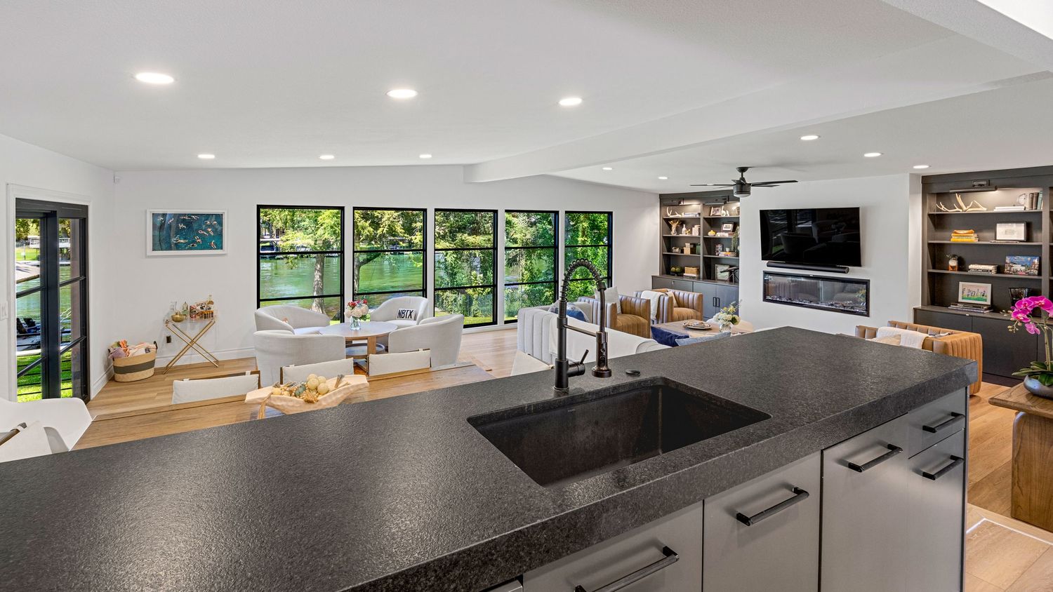 Modern open-concept kitchen with dark granite countertop and sink overlooking bright living room with large windows and built-in shelving.