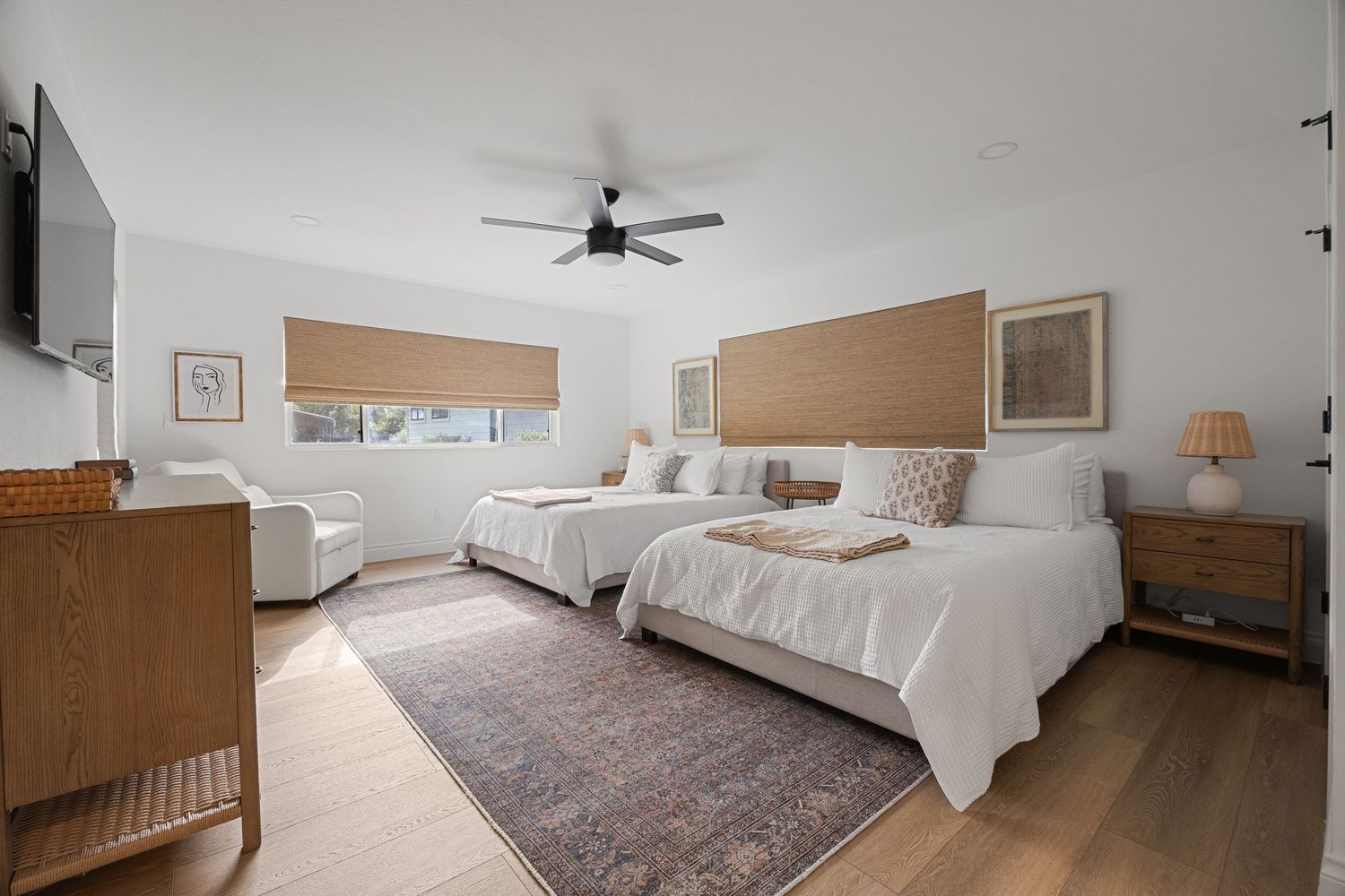 Bright guest bedroom featuring two queen beds with woven headboards, warm wood dresser, patterned area rug, and ceiling fan.