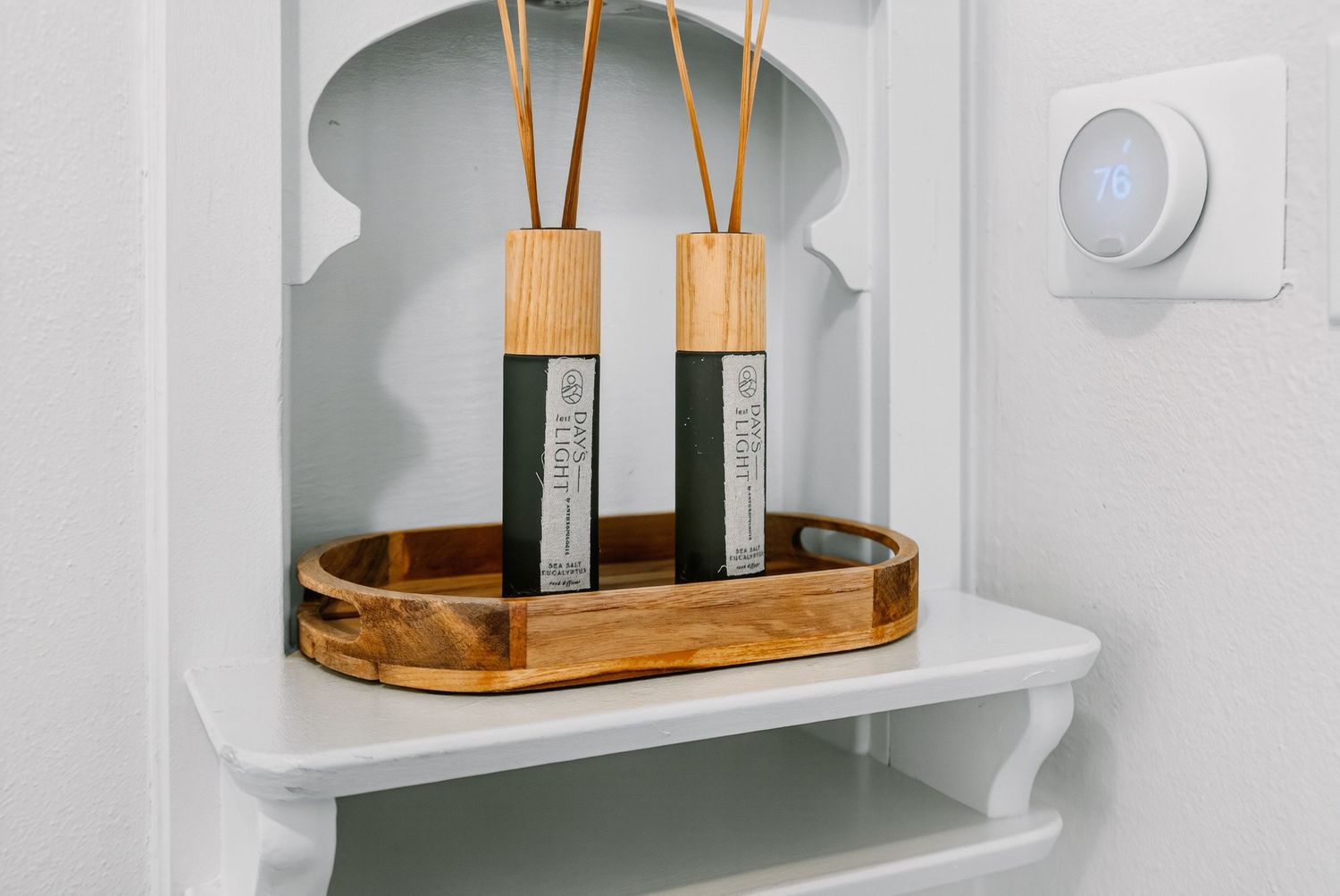 Reed diffusers with wooden sticks sit in a bamboo tray on a white bathroom shelf next to a motion sensor.