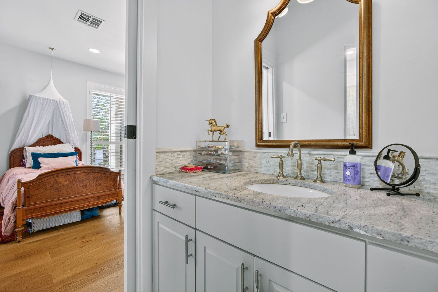 Elegant bathroom vanity with granite countertop, gold mirror, and white cabinets adjoining a charming bedroom with canopy bed.