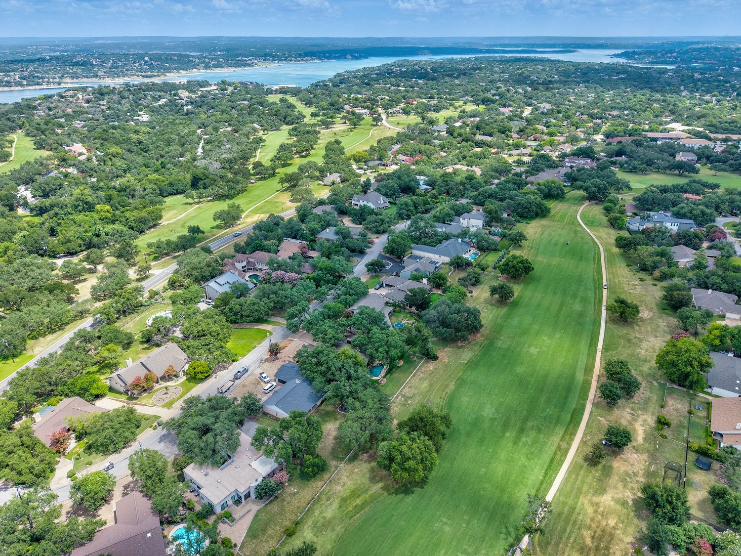 Aerial view of a lush Texas Hill Country neighborhood with golf course fairways, oak trees, and a lake visible in the background.