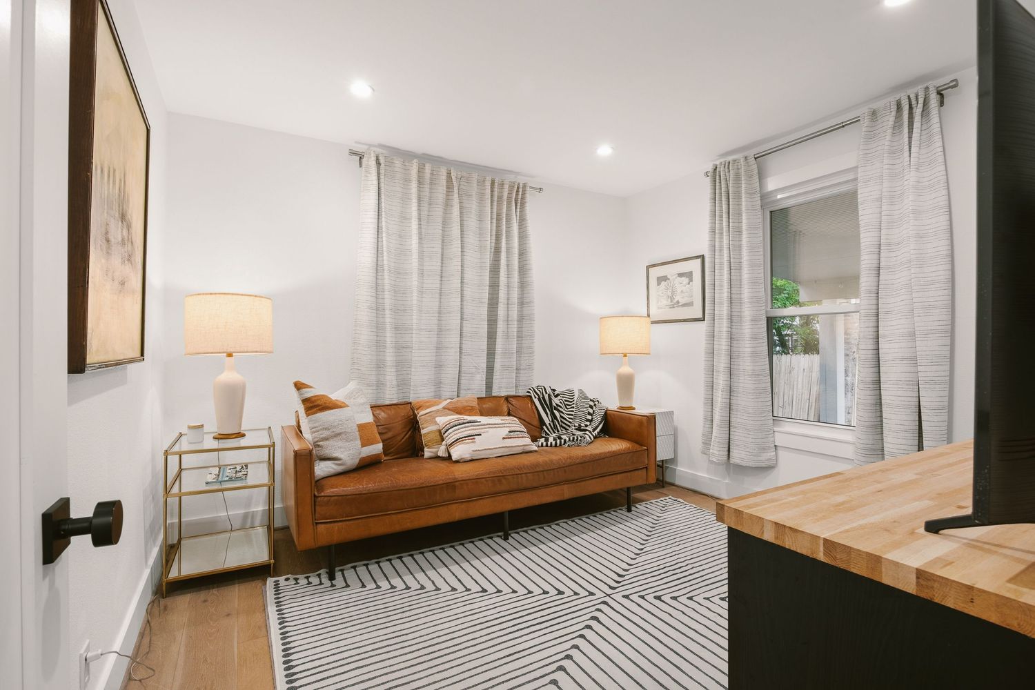 Cozy living room with brown leather sofa, geometric rug, white walls, and natural light from large windows with gray curtains.