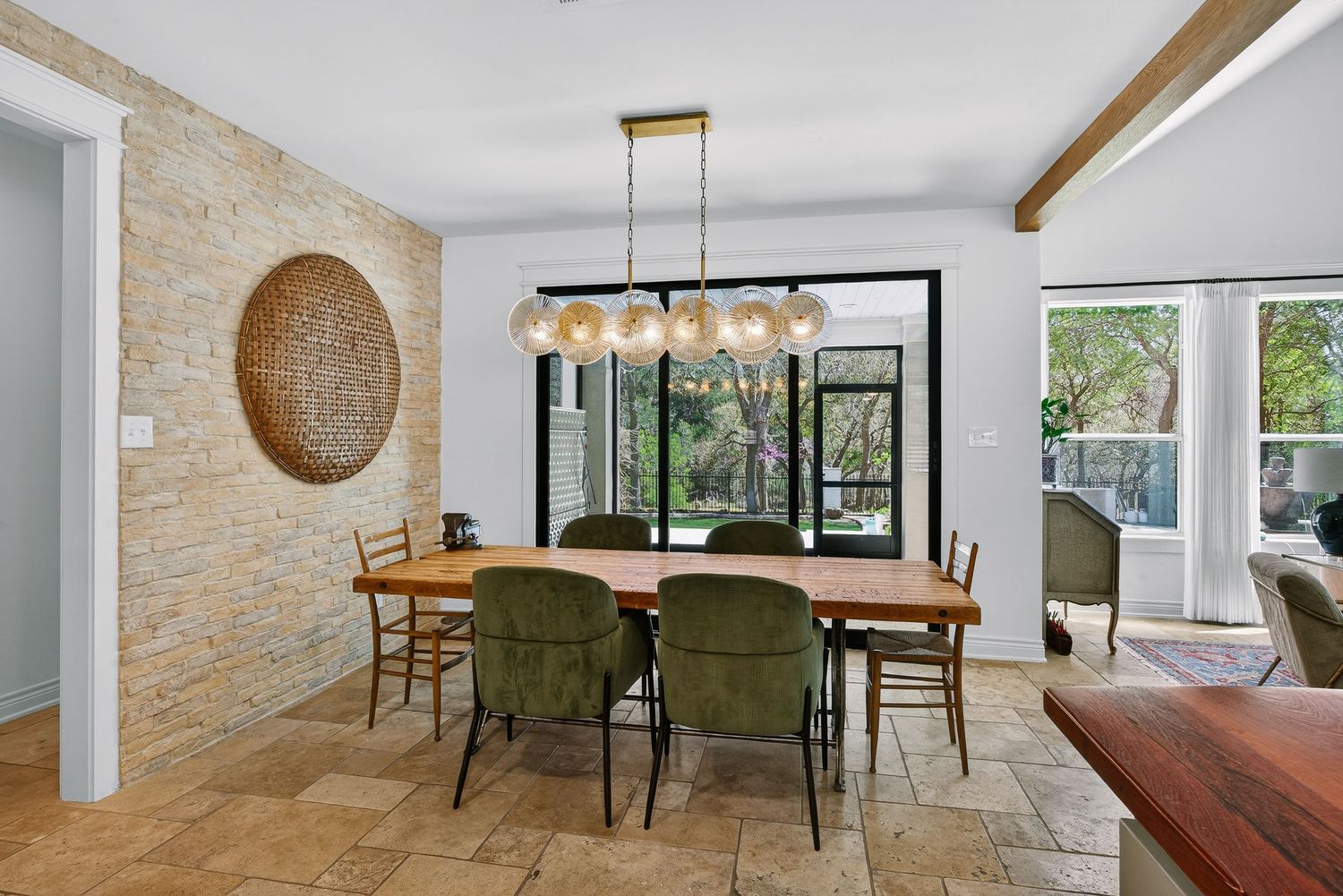 Modern dining room with wood table, green chairs, stone accent wall, and large glass doors overlooking lush greenery.