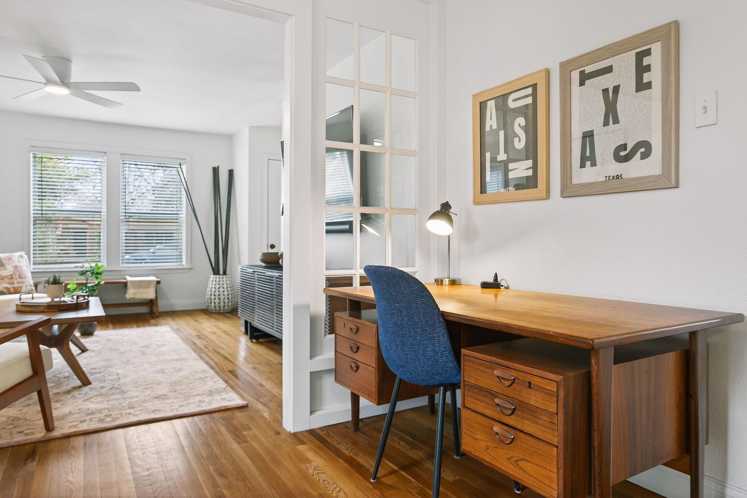 Modern home office nook with wooden desk and blue chair adjacent to bright living room with hardwood floors.