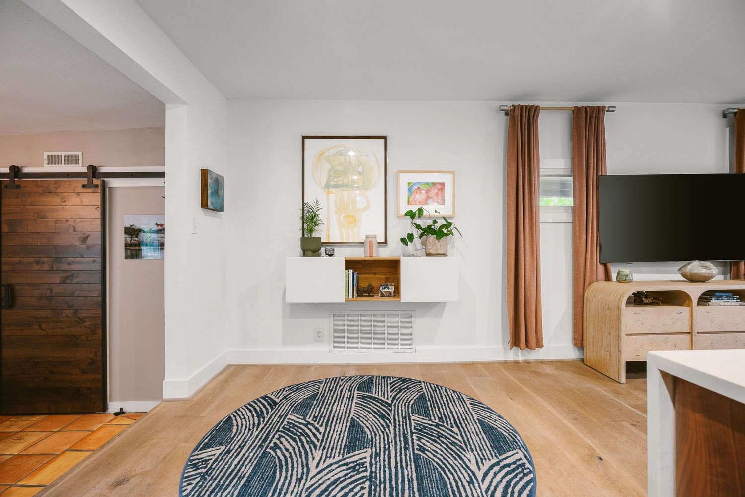 Bright living room with hardwood floors, barn door, round patterned rug, white walls, and orange curtains framing a window.