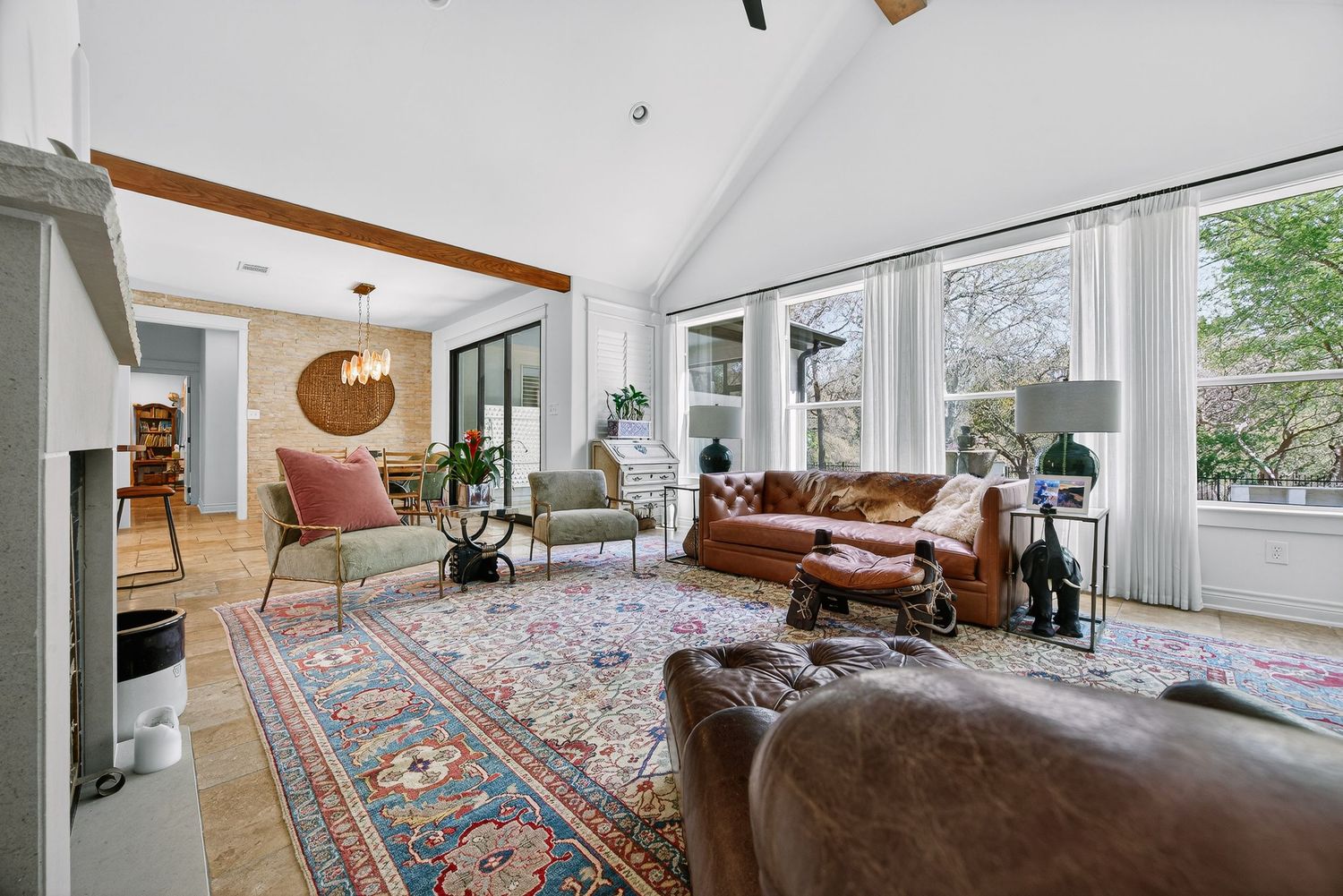 Bright living room with vaulted ceilings, leather sofas, a Persian rug, stone fireplace, and large windows overlooking lush greenery.