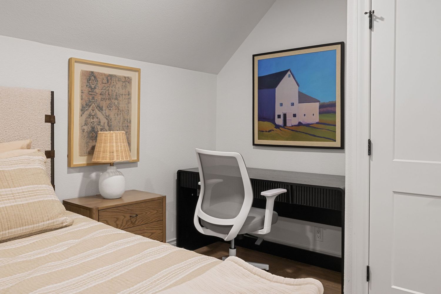 Cozy bedroom with striped bedding, wooden desk, white office chair, and colorful barn artwork on white walls.