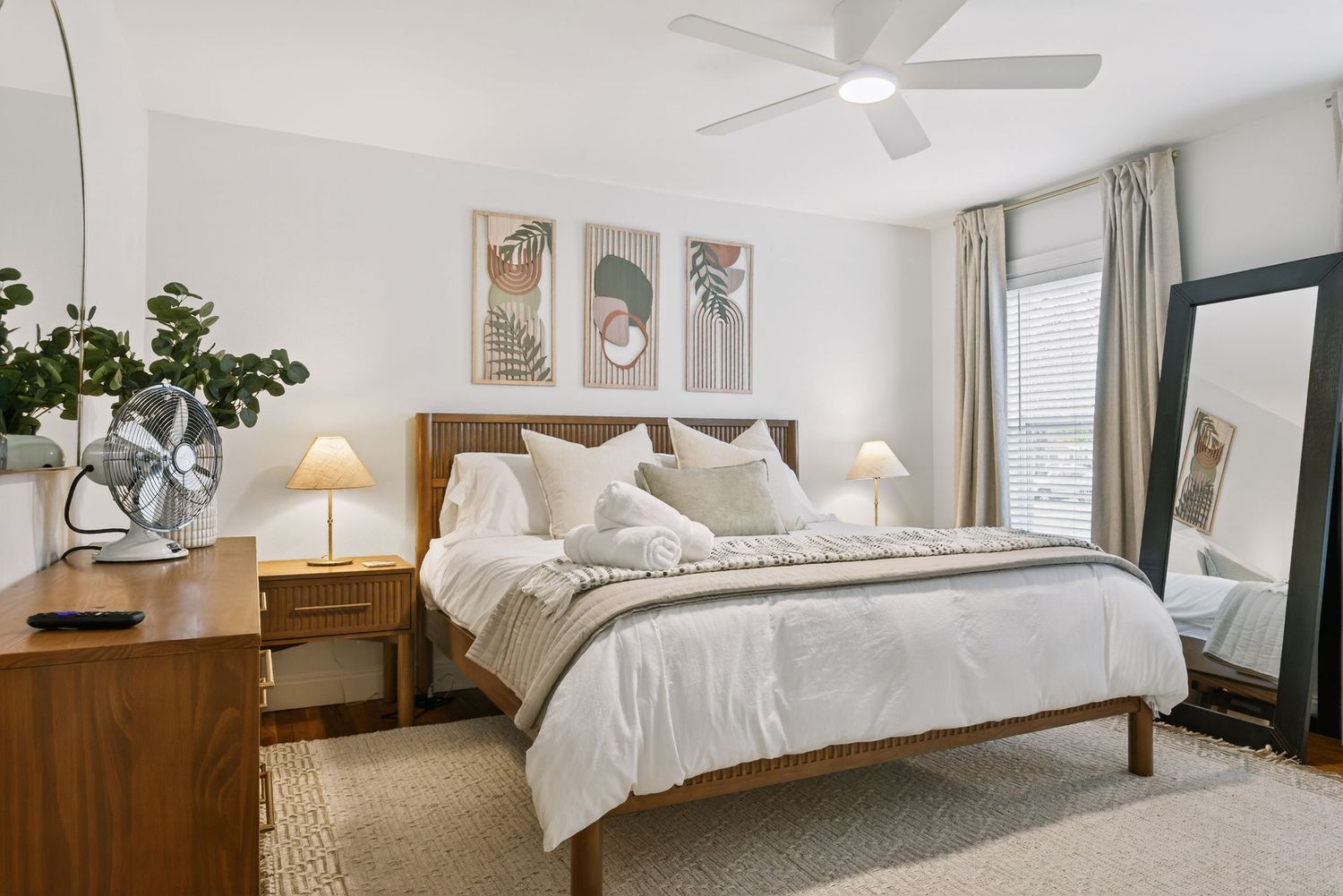 Bright and airy modern bedroom with white bedding, ceiling fan, and natural light streaming through windows.