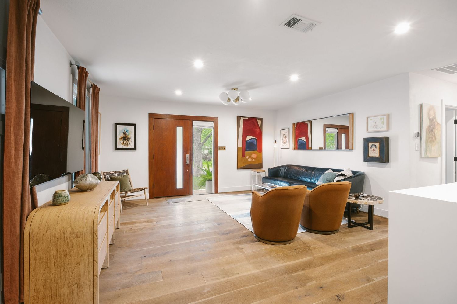 Bright living room with light hardwood floors, blue sofa, brown chairs, recessed lighting, and a wooden front door.