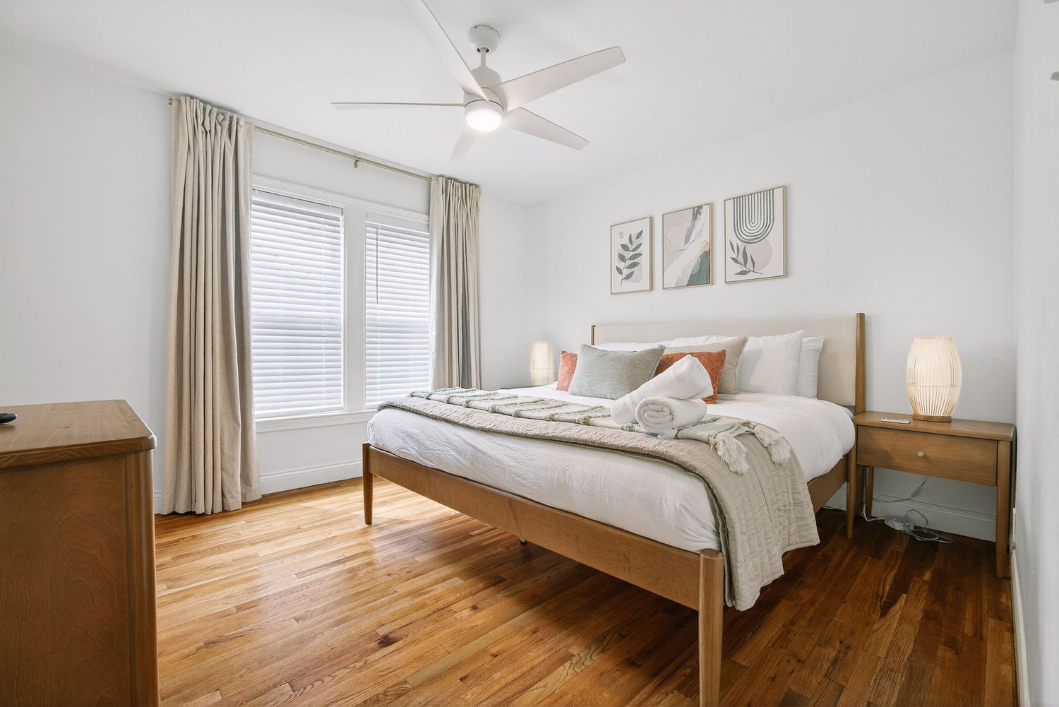 Modern bedroom with hardwood floors, white walls, beige curtains, and a wooden bed frame with white bedding.