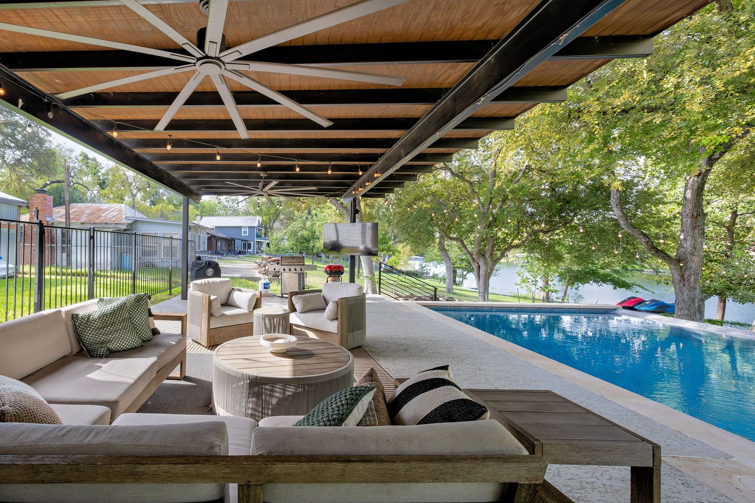 Covered outdoor patio with seating area, large ceiling fan, string lights, and sparkling swimming pool overlooking trees.